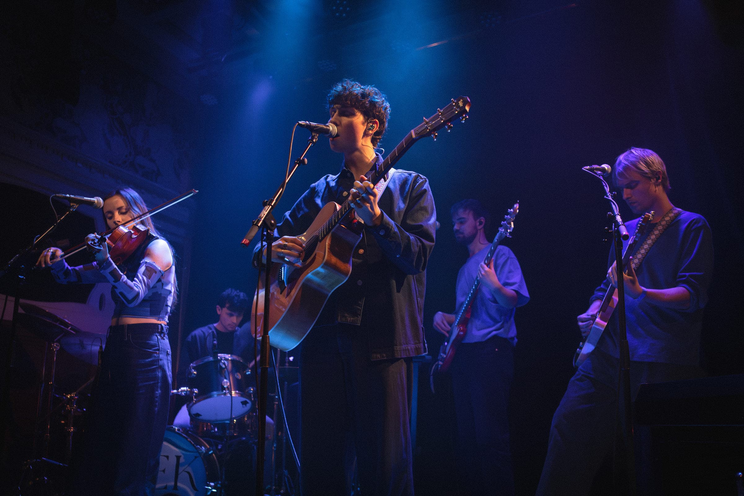 Concertfoto van Quinn Anderson: een band in blauw podiumlicht, de zanger met akoestische gitaar in het midden